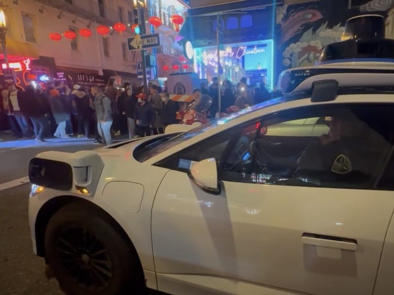 A sleek, autonomous Waymo white car paused at a bustling Chinatown intersection under the night sky, with people strolling and vibrant signs illuminating the background.